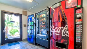 Une rangée de distributeurs automatiques aux couleurs de Pepsi et de Coca-Cola, une porte donnant sur un espace verdoyant et un sol carrelé.