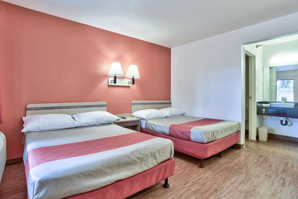 Une chambre d’hôtel avec deux lits, une table de chevet, une lampe, une poubelle et une salle de bain visible à travers une porte ouverte.