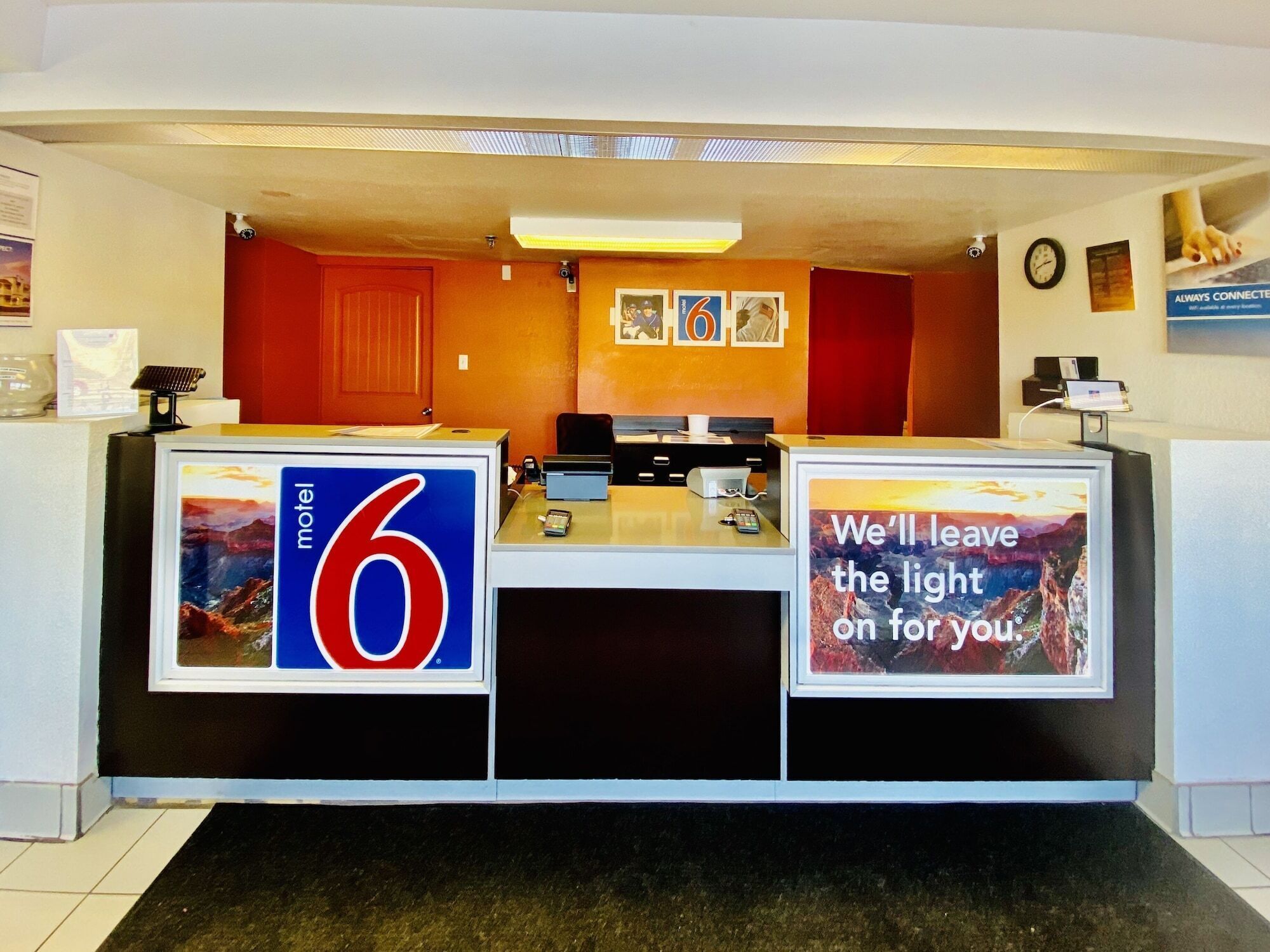 Een receptiebalie van een motel met een groot bord waarop '6' staat, een klok en een lijst met de tekst 'We houden het licht voor je aan'.