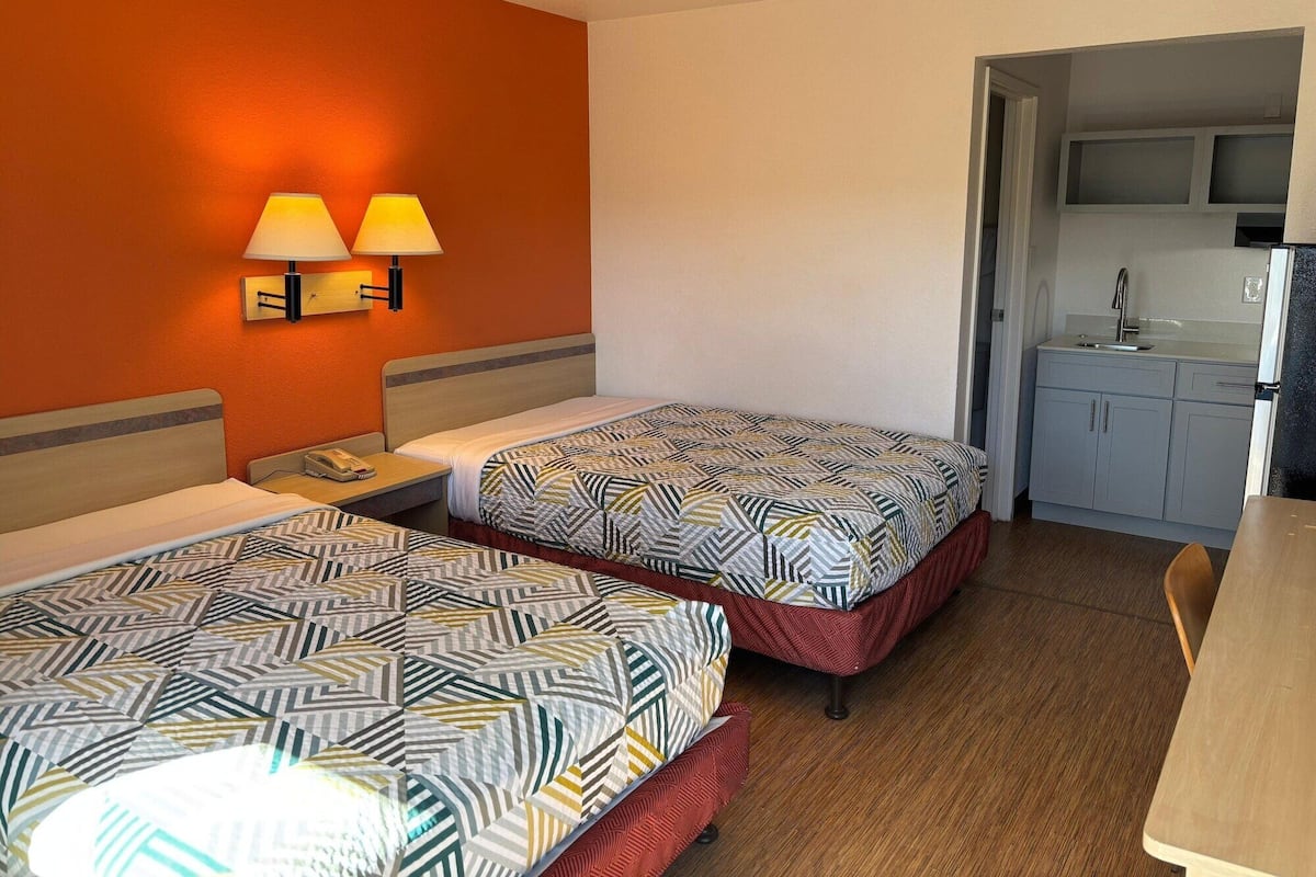 Une chambre d’hôtel avec deux lits, un petit bureau, un téléphone et une kitchenette équipée d’un évier et de placards.