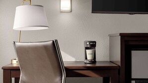 Quarto de hotel com uma poltrona de couro, mesa de cabeceira com cafeteira e TV fixada na parede.