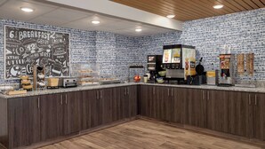 Balcão de pequeno-almoço com menu em quadro negro, máquinas de café e exposição de fruta.