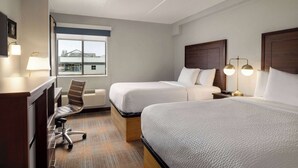 Quarto de hotel com duas camas, uma escrivaninha, uma cadeira e uma janela com vista para edifícios.