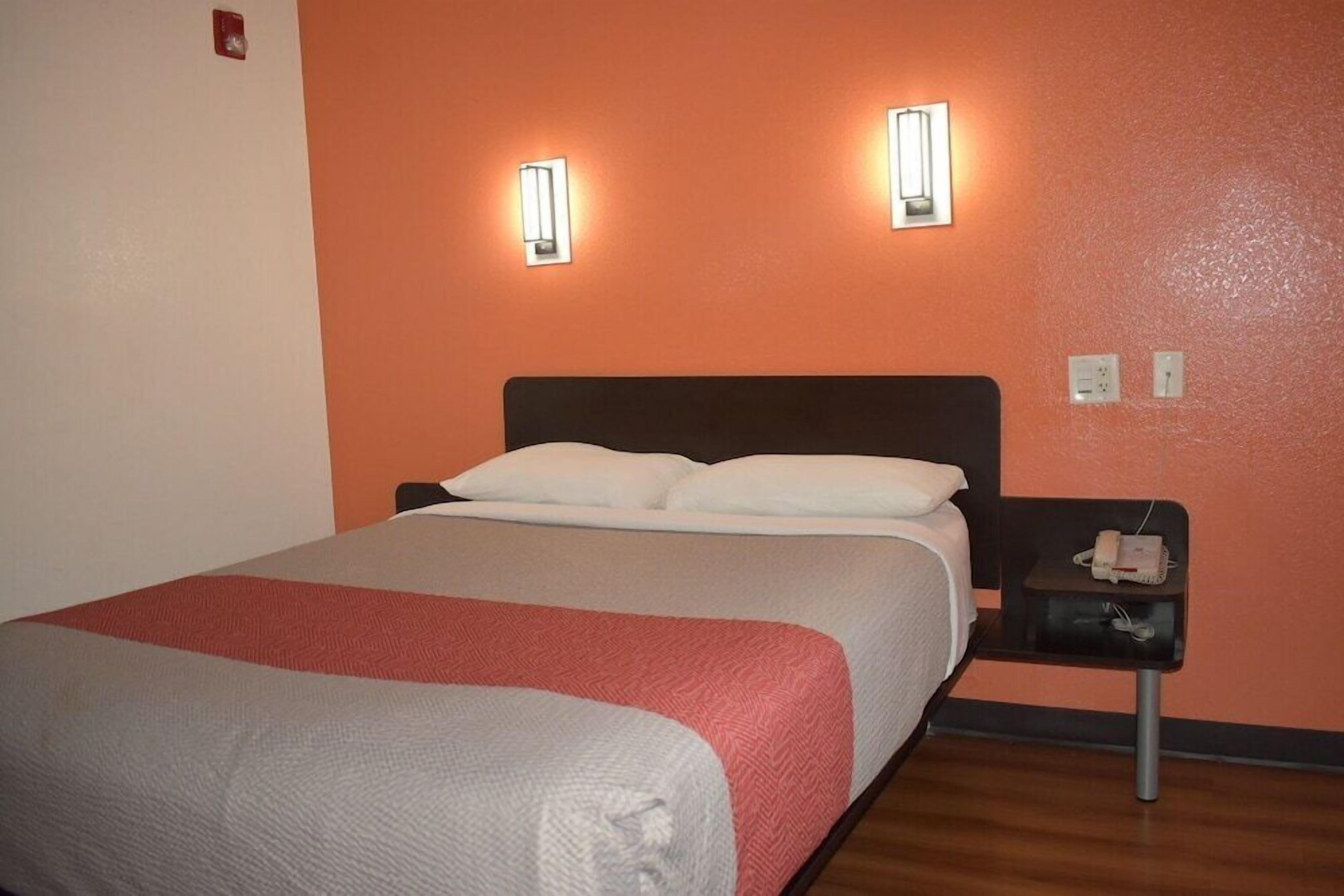 A hotel room with a bed, two wall-mounted lights, a bedside table with a phone, and a red and white bedspread.