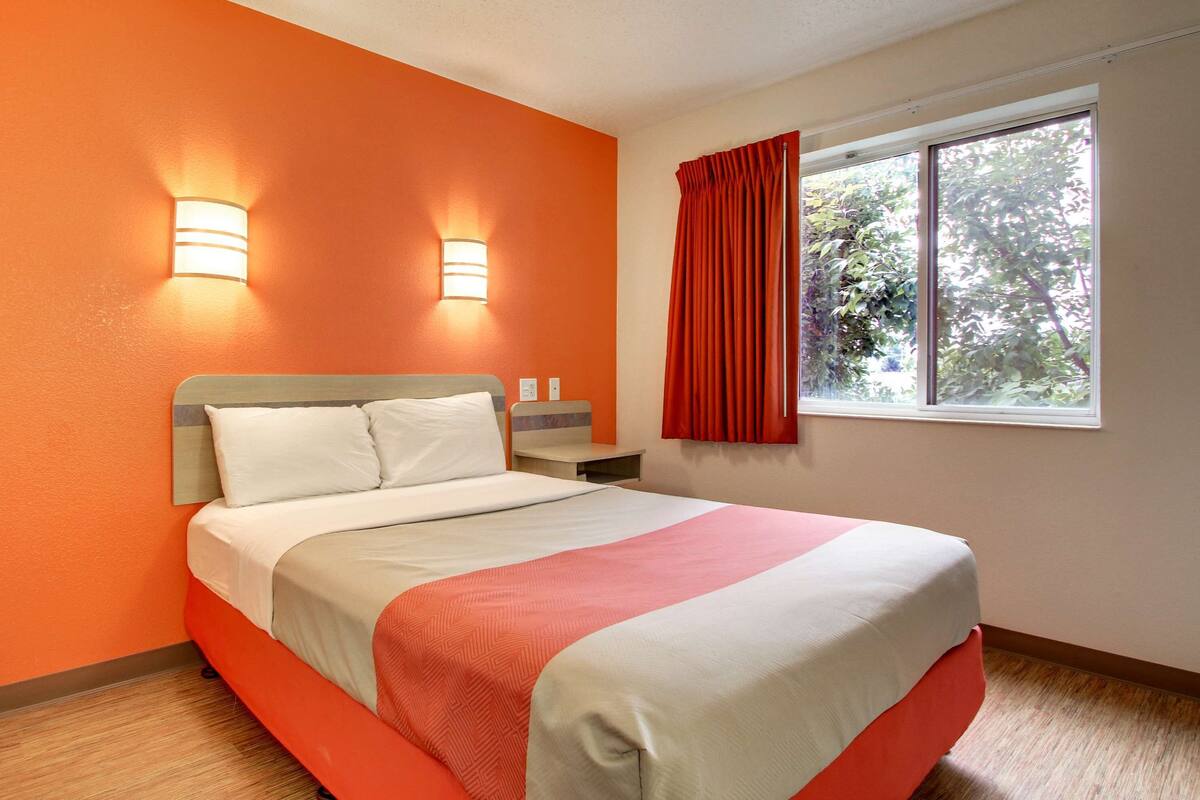 A hotel room with a bed, two wall-mounted lights, a window with red curtains, and a view of greenery outside.