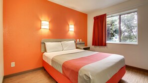 A hotel room with a bed, two wall-mounted lights, a window with red curtains, and a view of greenery outside.