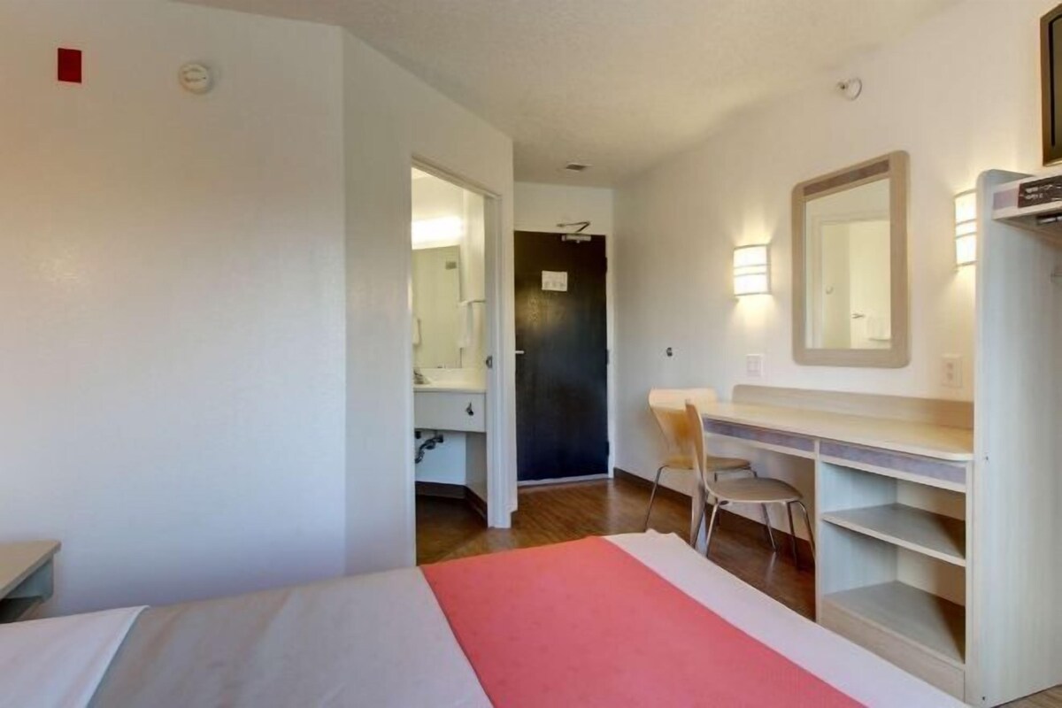 A hotel room with a bed, a desk with a chair, a mirror, and a shelf.