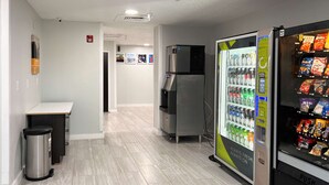 A clean, modern hallway with a vending machine stocked with beverages and snacks, a trash bin, and a refrigerator. - Motel 6 Springfield, IL (Springfield)