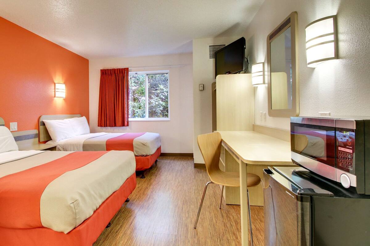 A hotel room with two beds, a desk, a TV, and a window with curtains.