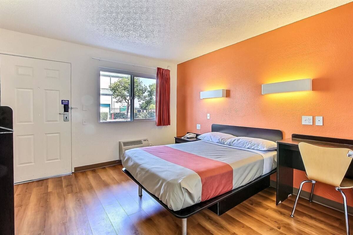 A hotel room with a bed, a desk, a chair, a window, and a door.