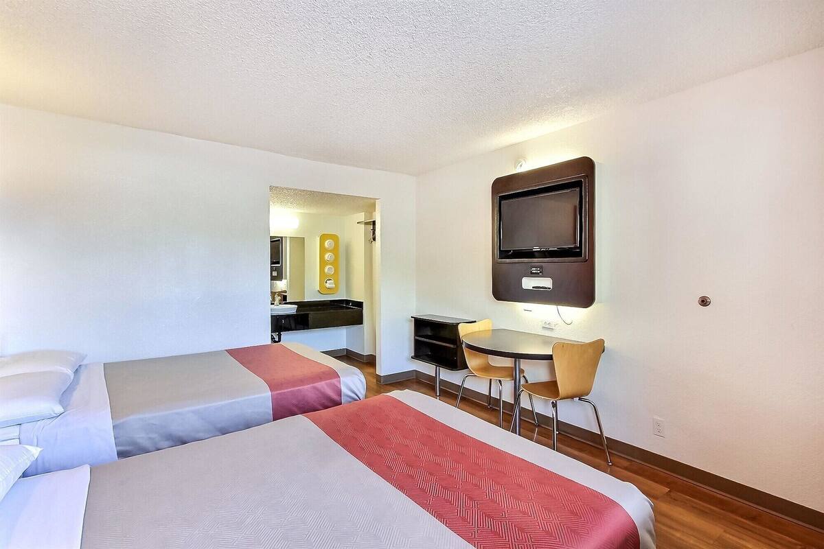 A hotel room with two beds, a desk with a chair, a TV, and a bathroom visible through an open door.