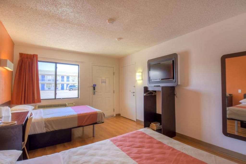 A hotel room with two beds, a television, a window with curtains, and a view of the street.