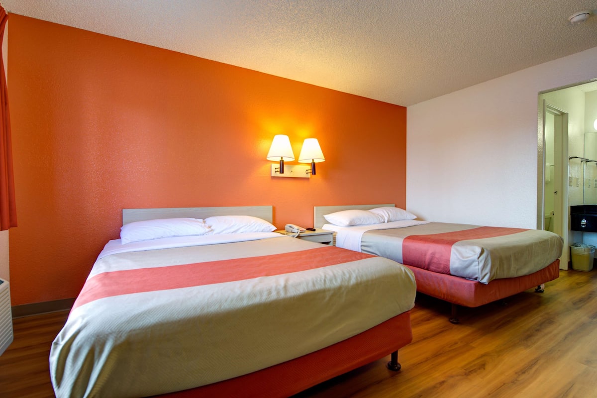 A hotel room with two beds, a nightstand, a lamp, and a bathroom visible through an open door.