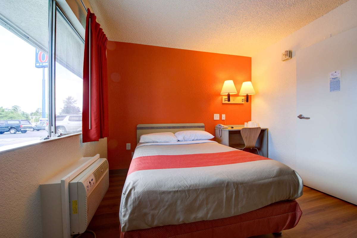 A hotel room with a large bed, a window with red curtains, a heater, and two wall-mounted lamps.