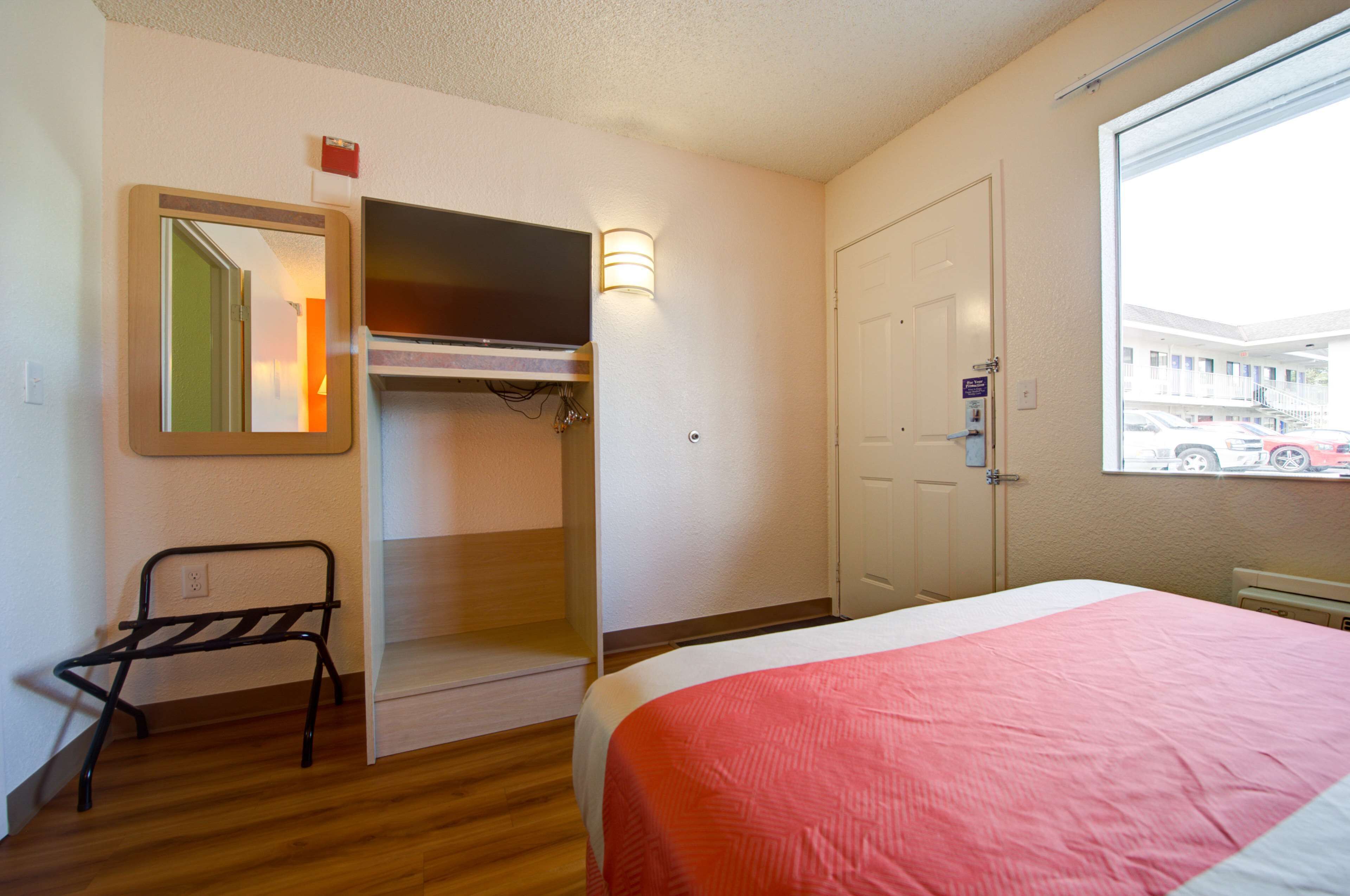 A hotel room with a bed, a television, a mirror, a small desk, and a window with a view of parked cars.