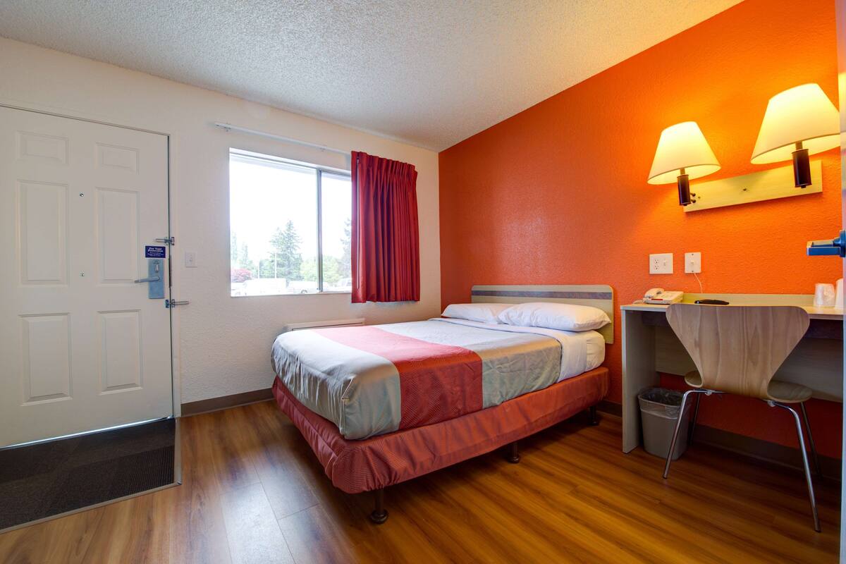 A hotel room with a bed, a desk, a chair, a window with curtains, and a door.