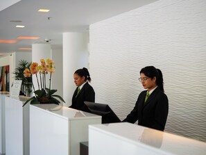 A reception desk with two receptionists in business attire, a potted plant with yellow flowers, and a textured wall in the background. - NM Lima Hotel (Lima)