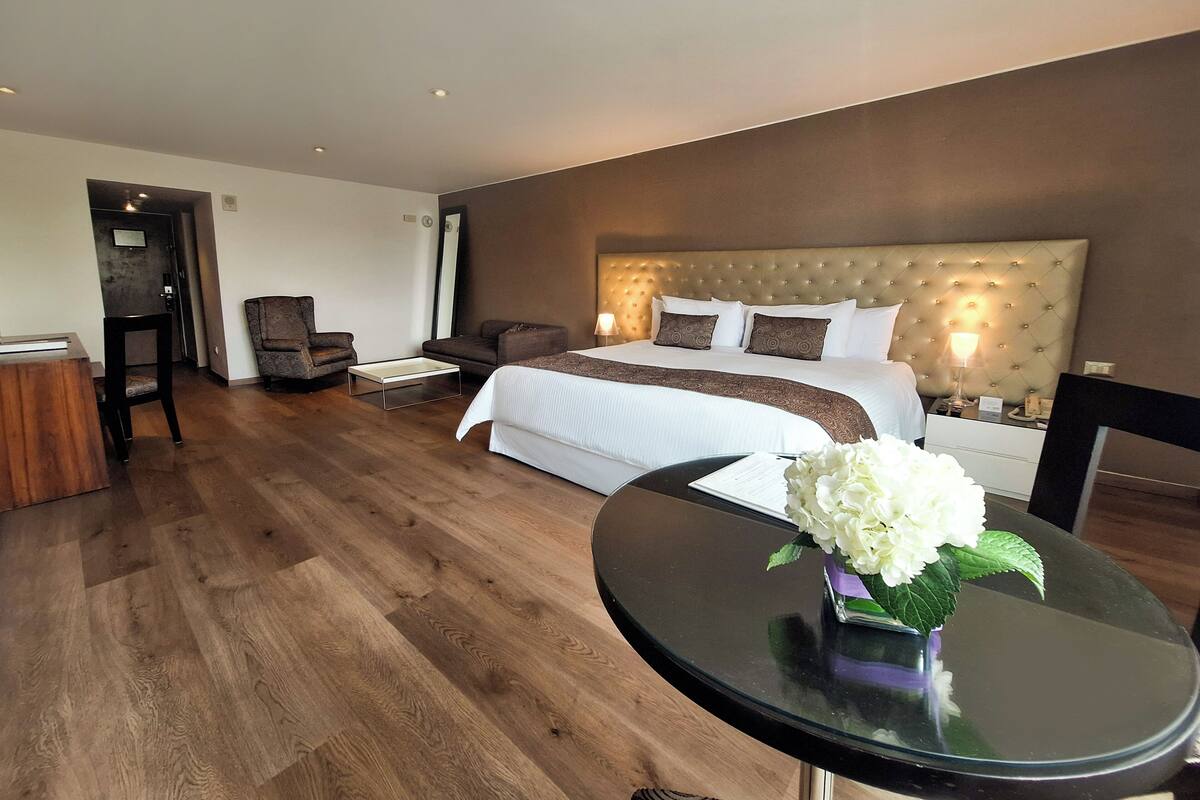 A hotel room with a large bed, a sitting area with a chair and a small table, and a glass table with a vase of flowers.