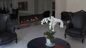 A modern lobby with a reflective floor, black upholstered chairs, a round table with a white orchid, and a fireplace with visible flames. - NM Lima Hotel (Lima)