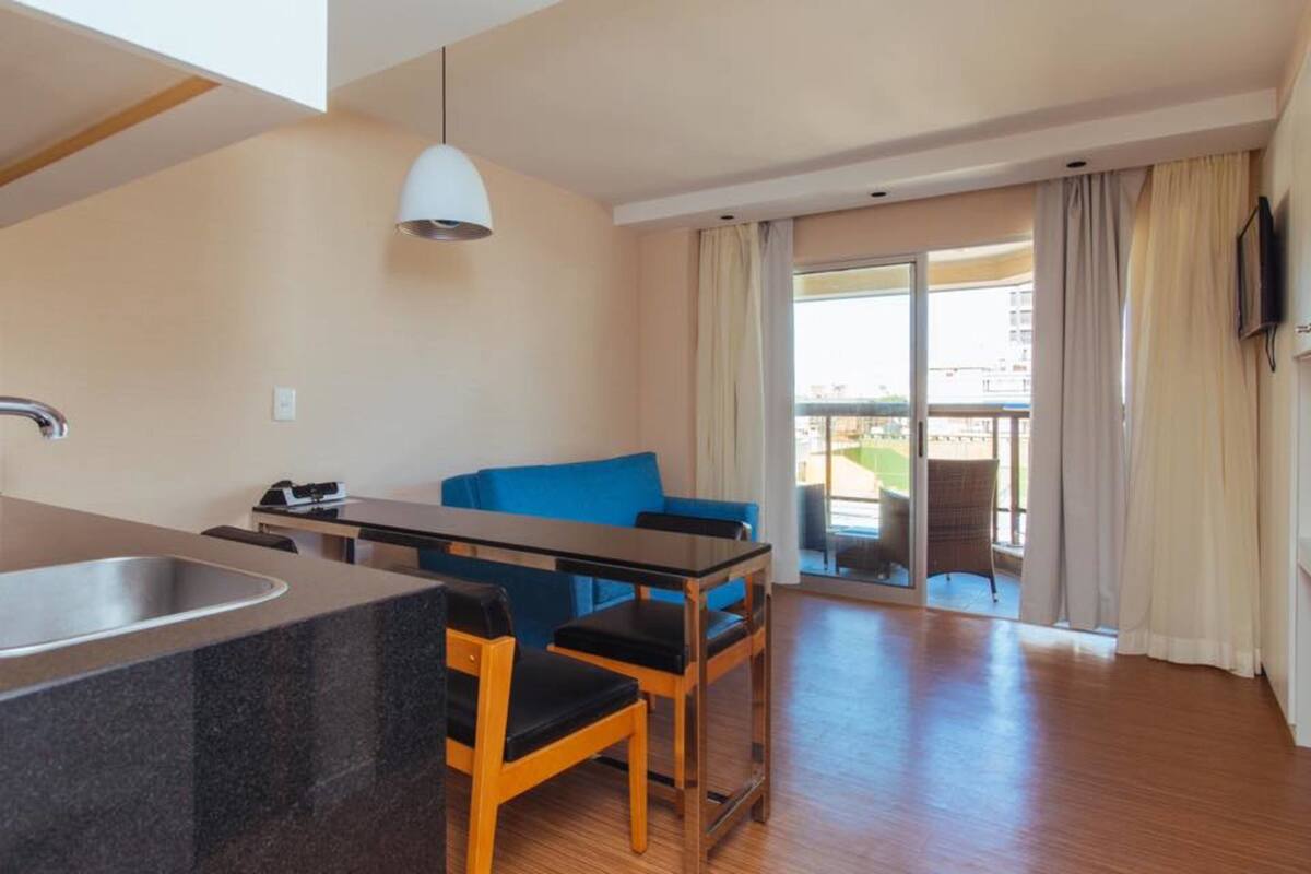 A modern hotel room with a kitchen area, a dining table with chairs, a blue sofa, and a balcony with curtains.