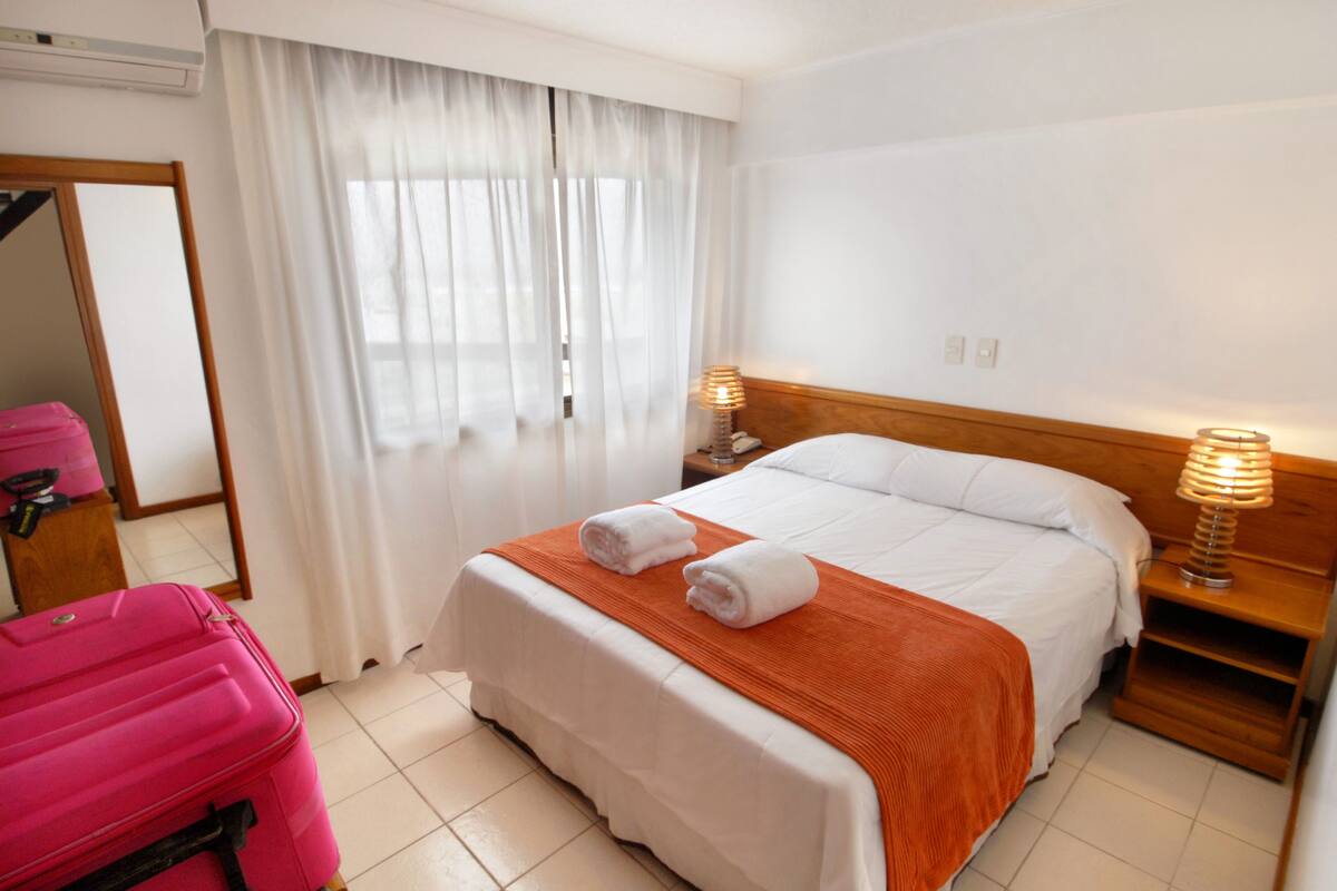 Una habitación de hotel con una cama, dos lámparas de noche, una mesita de noche de madera, un espejo, una maleta rosa y una ventana con cortinas blancas.