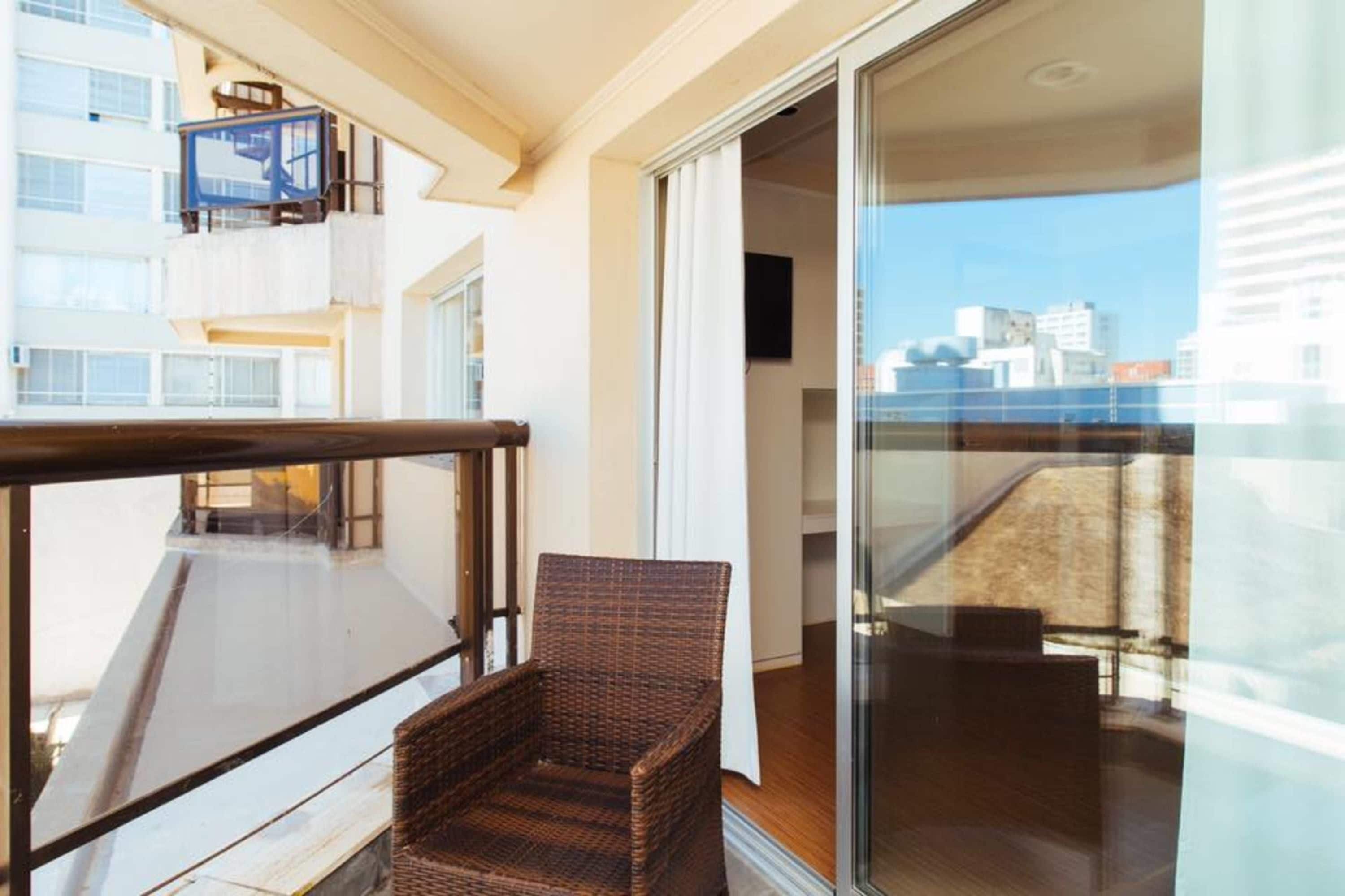 Ein Balkon mit Korbstuhl, einer Glastür mit Blick auf die Stadt und einer blauen Sesselbahn links.