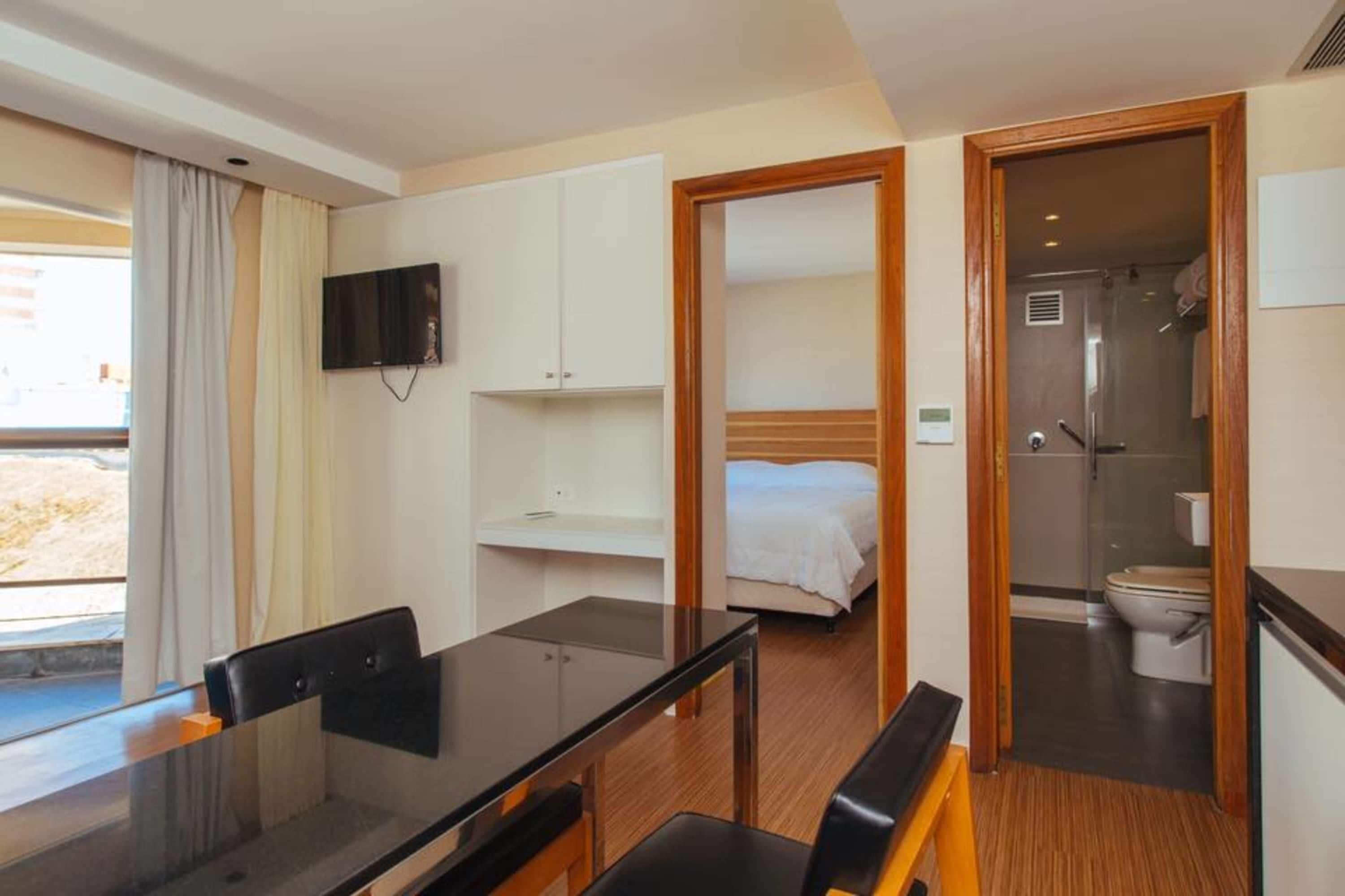 A compact hotel room with a bed, a small bathroom, a TV mounted on the wall, and a dining area with a table and chairs.