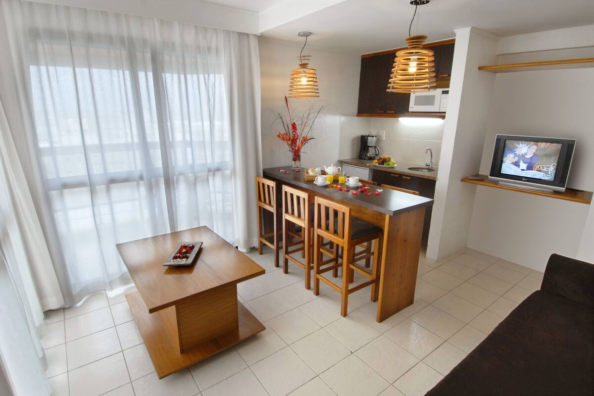 A modern living room with a wooden dining table, a sofa, a kitchen area with a microwave, and a TV.
