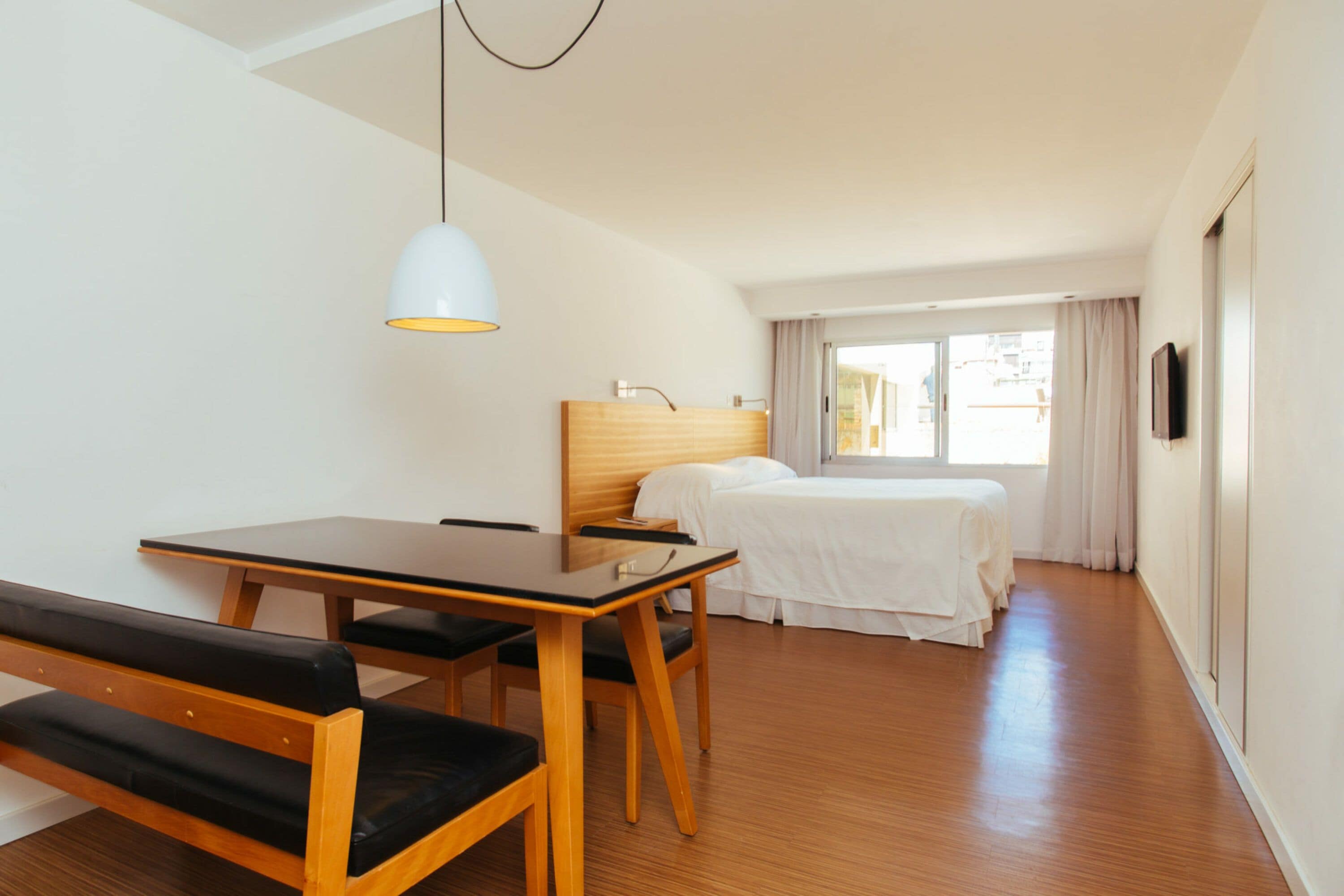 Una habitación de hotel con una mesa de madera y un banco, una cama con ropa blanca, un cabecero y un televisor en la pared.