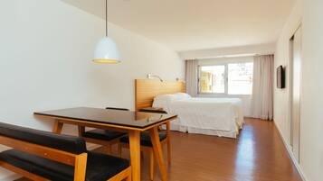Una habitación de hotel con una mesa de madera y un banco, una cama con ropa blanca, un cabecero y un televisor en la pared.