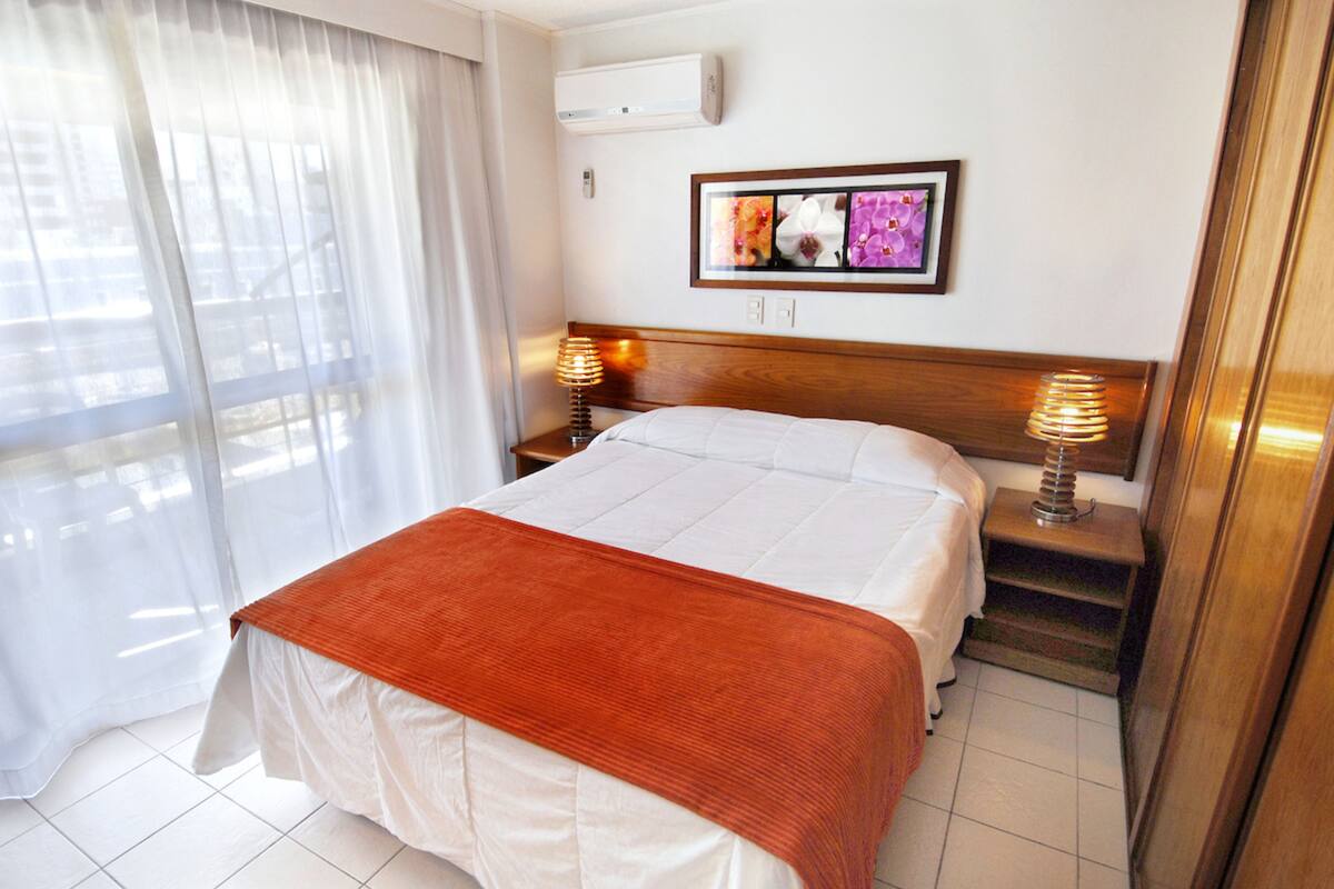 A hotel room with a large bed, two bedside tables with lamps, a framed picture on the wall, and a window with sheer curtains.