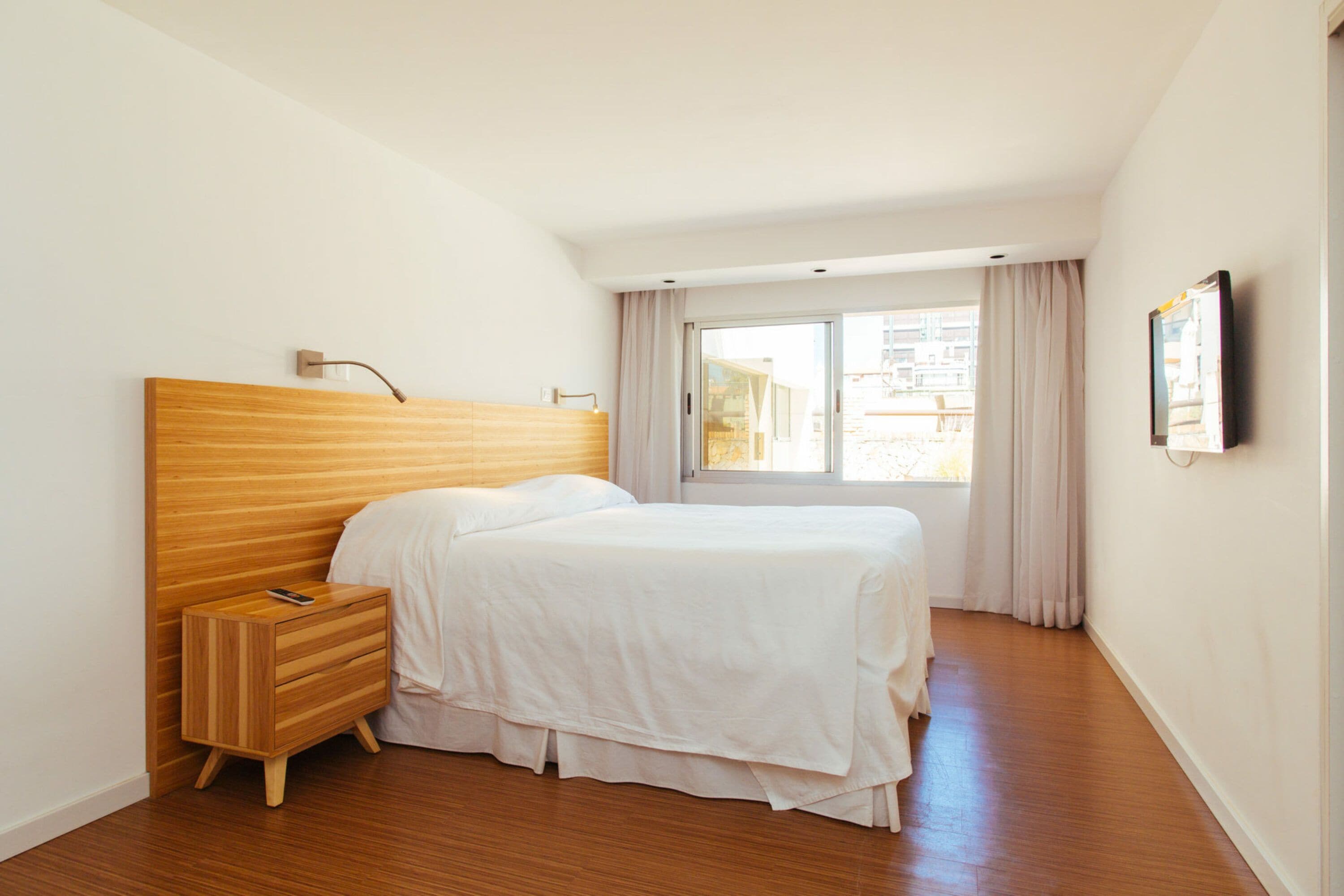 A bedroom with a wooden headboard, a single bed, a nightstand, a window with curtains, and a wall-mounted TV.