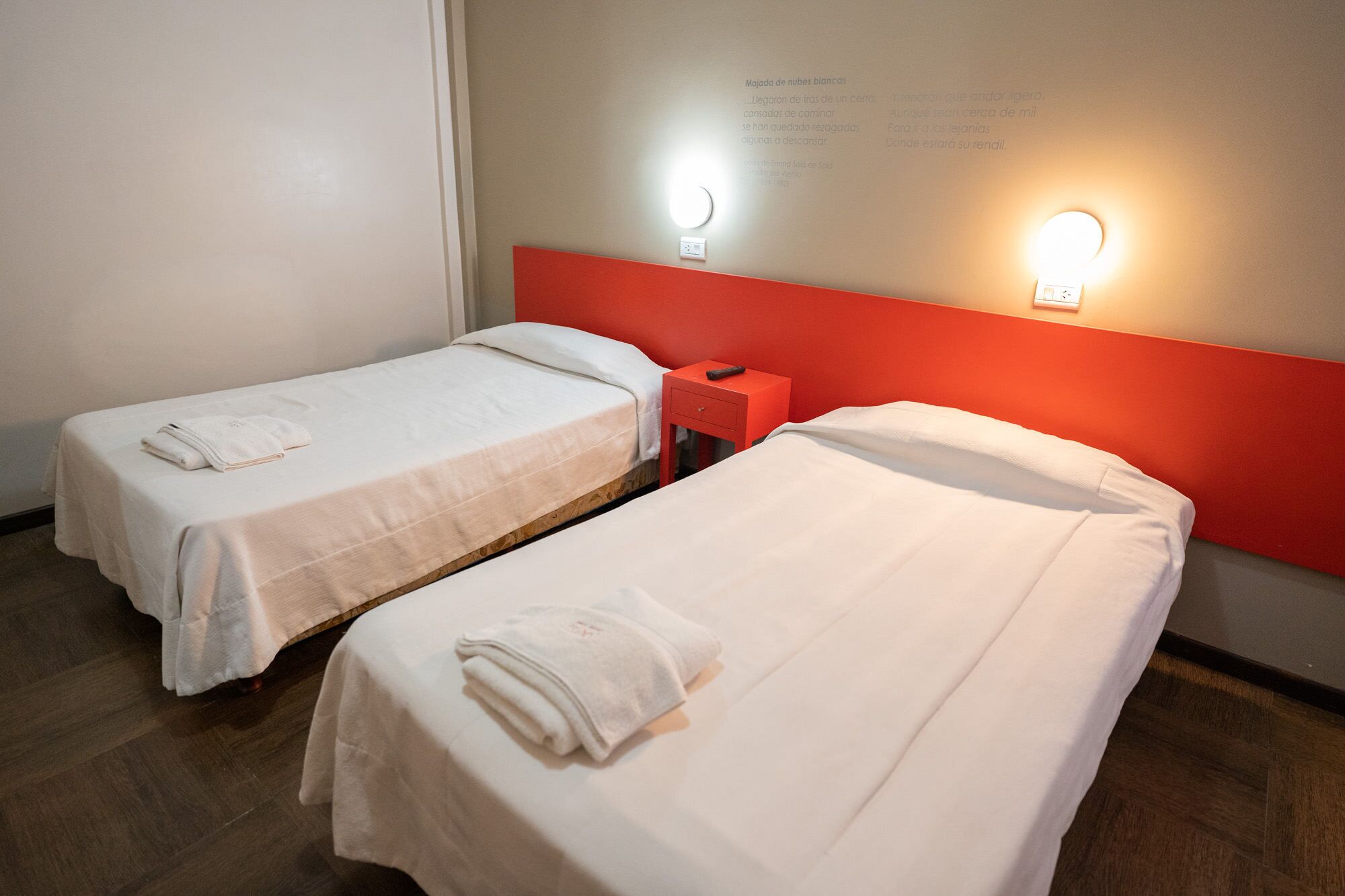 Two single beds in a room with a red headboard, white bedding, and a small bedside table with a remote control.