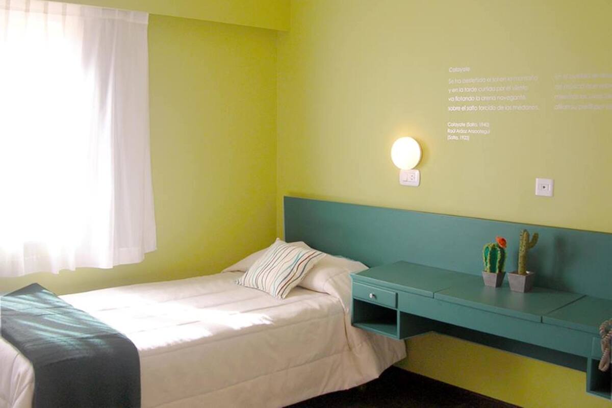 A room with a bed, a bedside table with a lamp, and a wall-mounted shelf with plants.