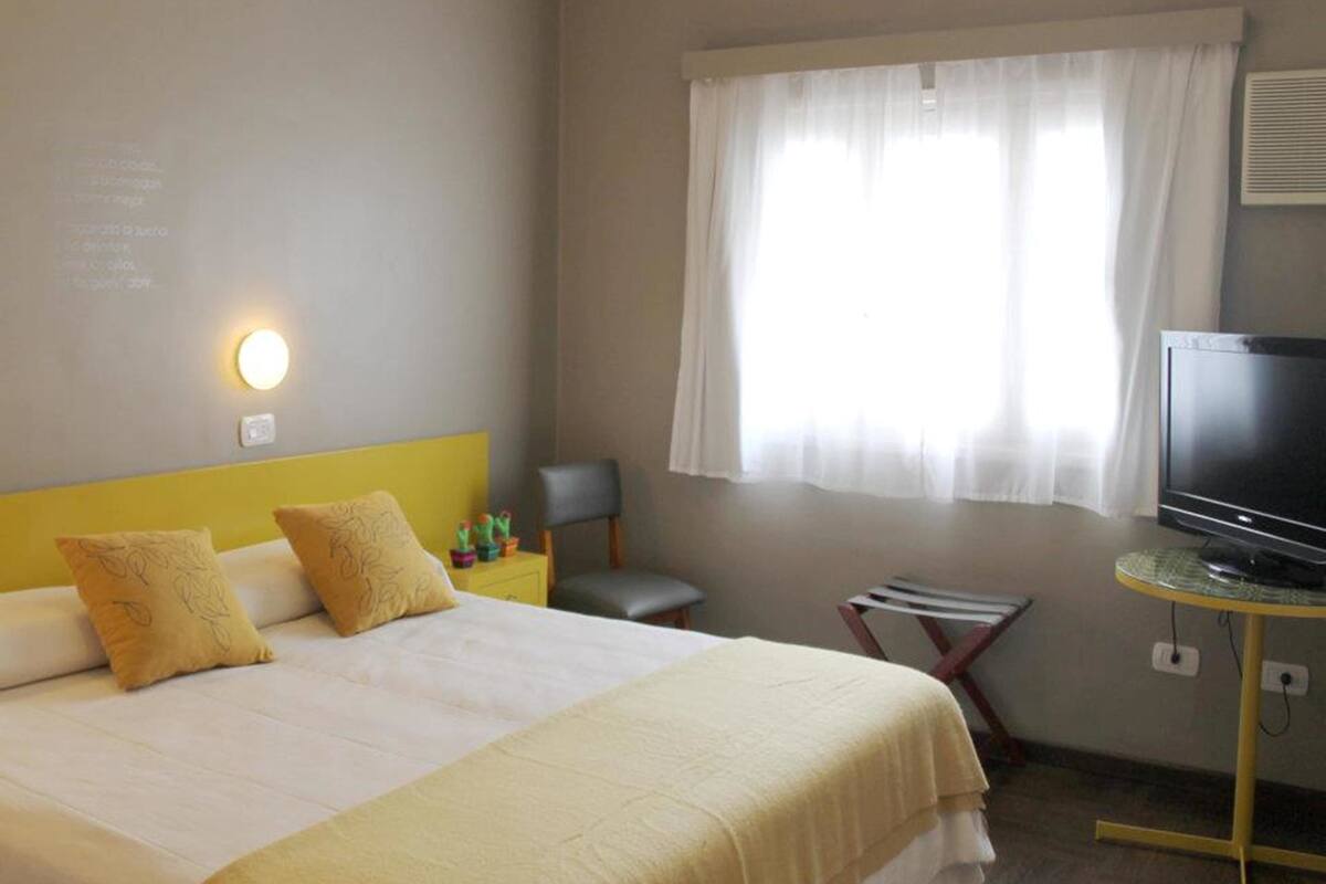 A hotel room with a bed, a television on a stand, a small table, and a window with curtains.