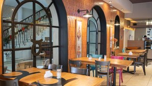 Ein Restaurant mit Holztischen und -stühlen, großen Fenstern mit Verzierungen und einem Küchenbereich im Hintergrund.