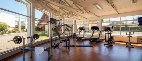 Academia com diversos equipamentos de exercícios, grandes janelas e vista para o exterior.