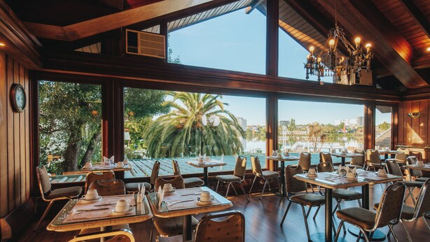 A restaurant with wooden interiors, large windows, and a view of palm trees and buildings. - Hotel Marina Villa del Rio (Valdivia)