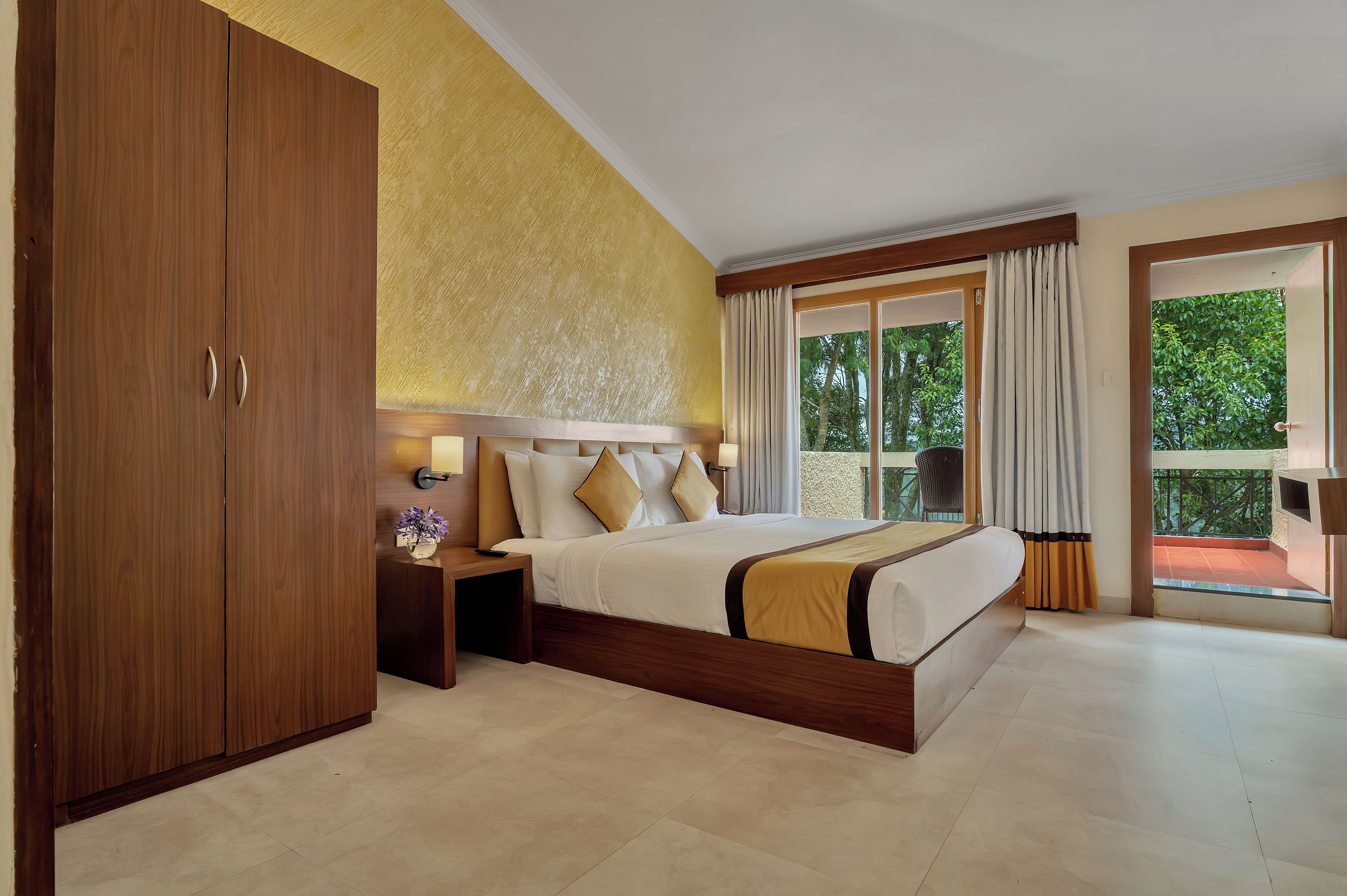 Een hotelkamer met een groot bed, een kledingkast, een nachtkastje met een lamp en uitzicht op groen door het raam.