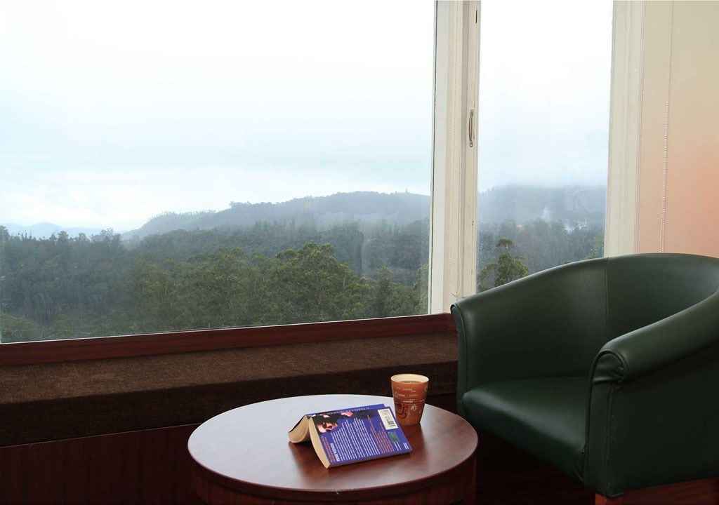 A room with a view of a forested landscape through a window, a round wooden table with a book and a cup, and a green leather chair.