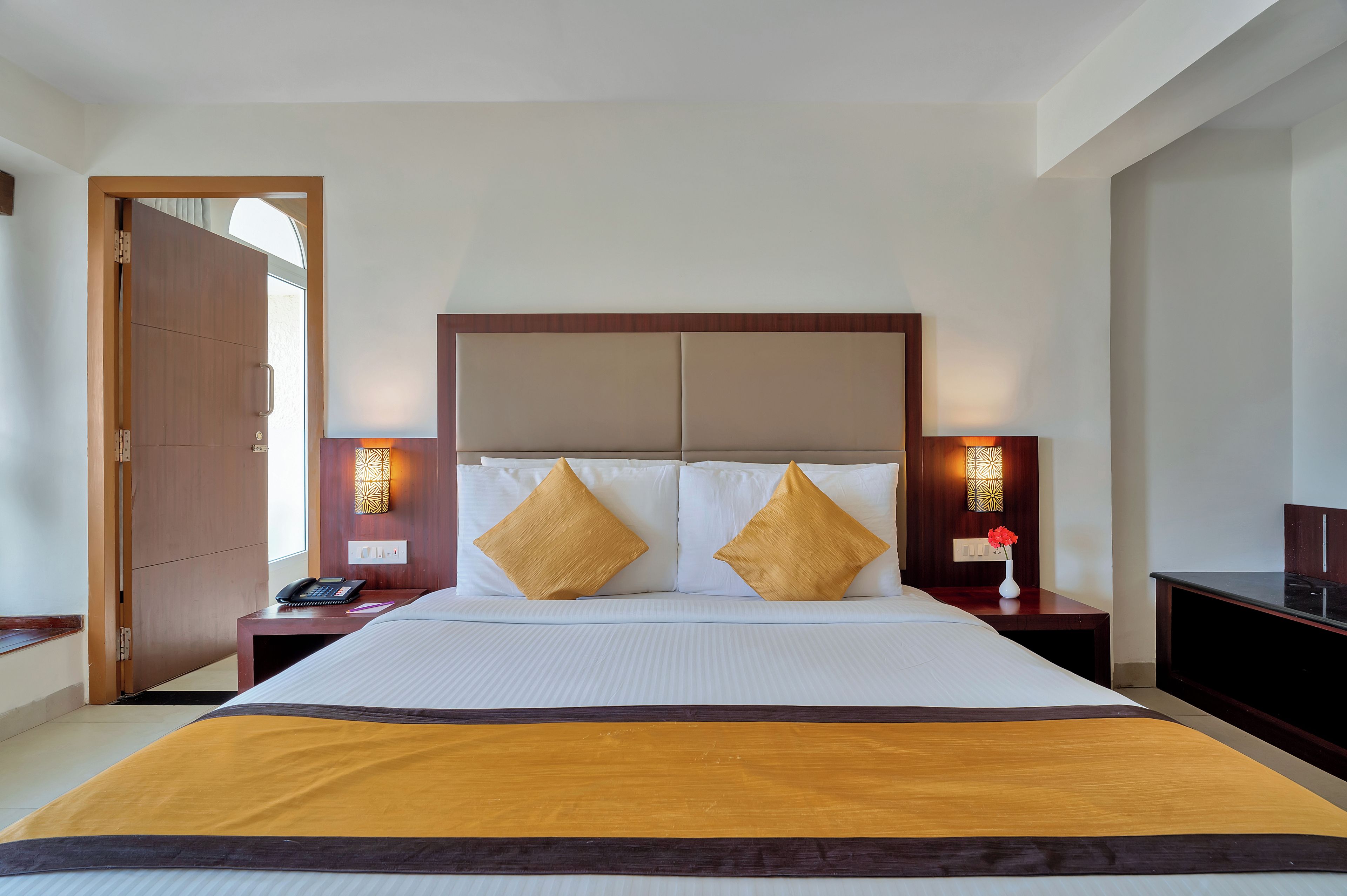 Une chambre d’hôtel avec un grand lit, deux tables de chevet, une petite table d’appoint, une porte en bois et un luminaire fixé au mur.