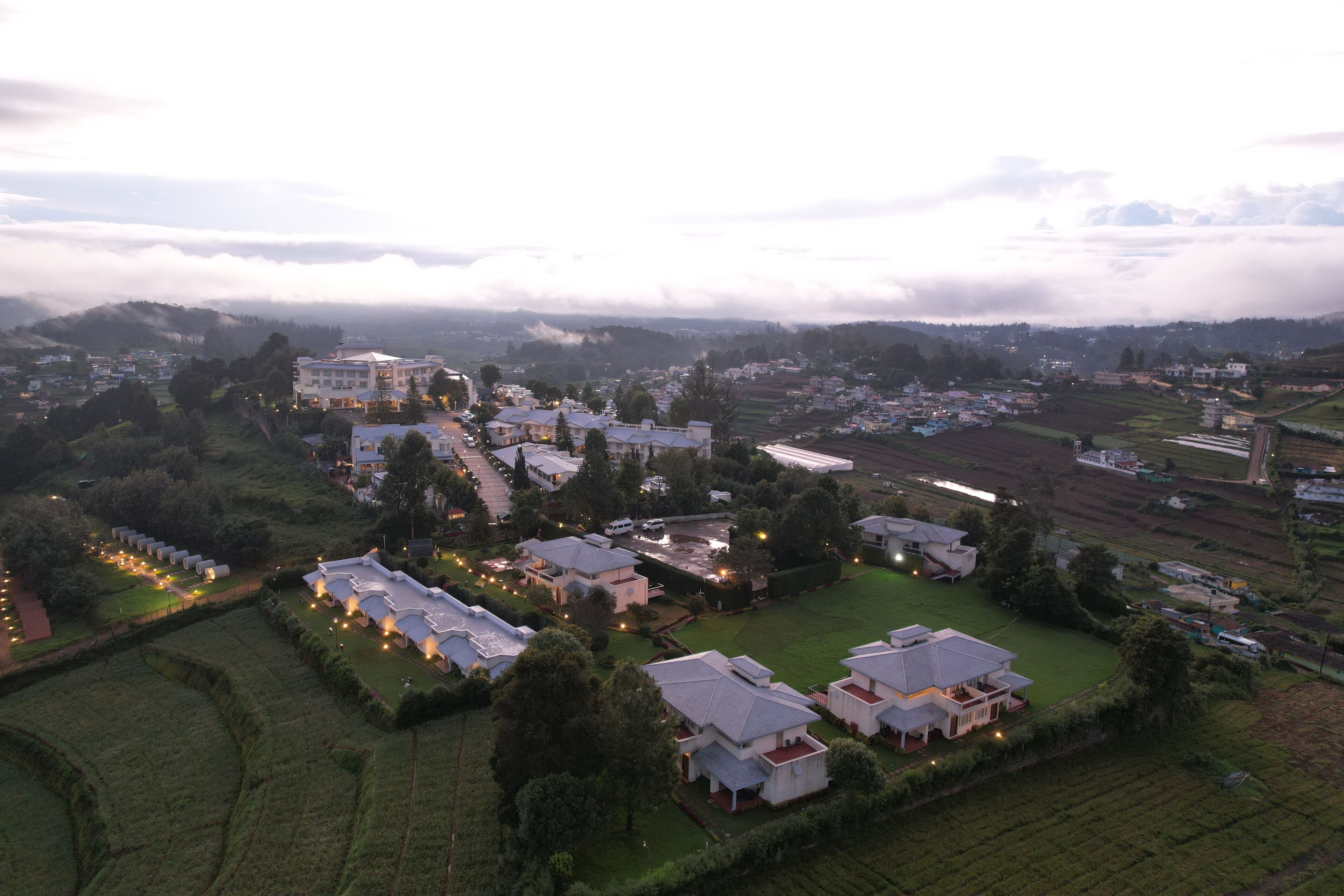 Vista aérea de una zona residencial con varias casas y un edificio grande, rodeada de campos verdes y un paisaje montañoso.