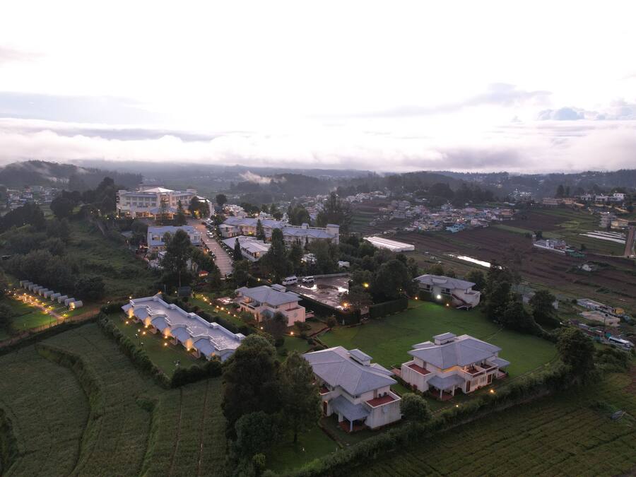 Vista aérea de una zona residencial con varias casas y un edificio grande, rodeada de campos verdes y un paisaje montañoso.