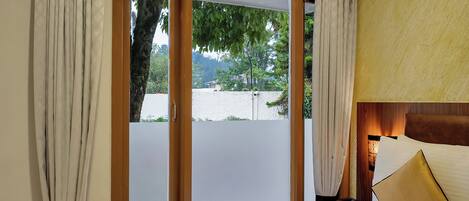 A hotel room with a bed, a bedside table, a lamp, and a view through a sliding door to the outdoors.