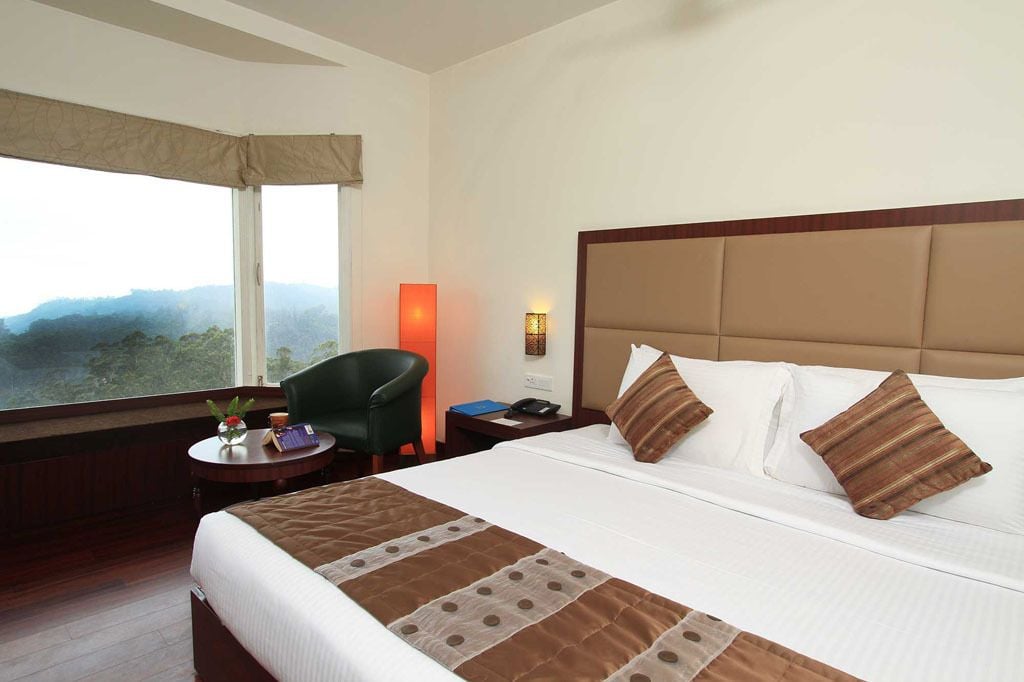 Habitación de hotel con una cama grande, una mesita de noche, una silla, una lámpara y vistas a la montaña.