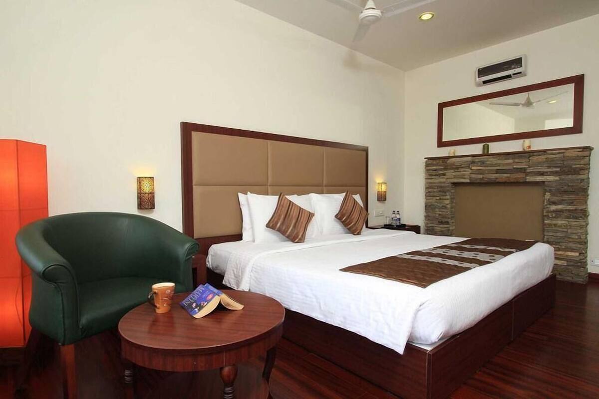 Une chambre d’hôtel avec un grand lit, un fauteuil vert, une petite table ronde, une cheminée et un ventilateur de plafond.