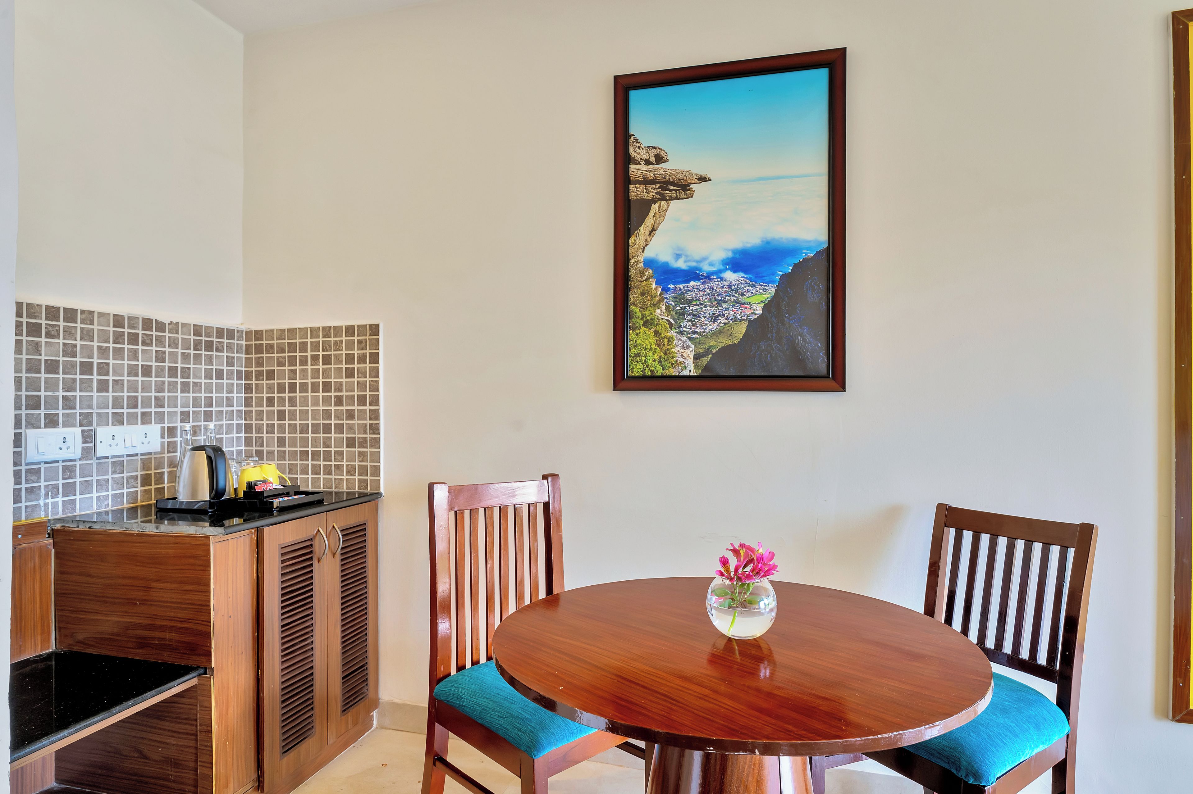 A dining area with a round table, two chairs, a vase with flowers, and a painting of a coastal landscape on the wall.