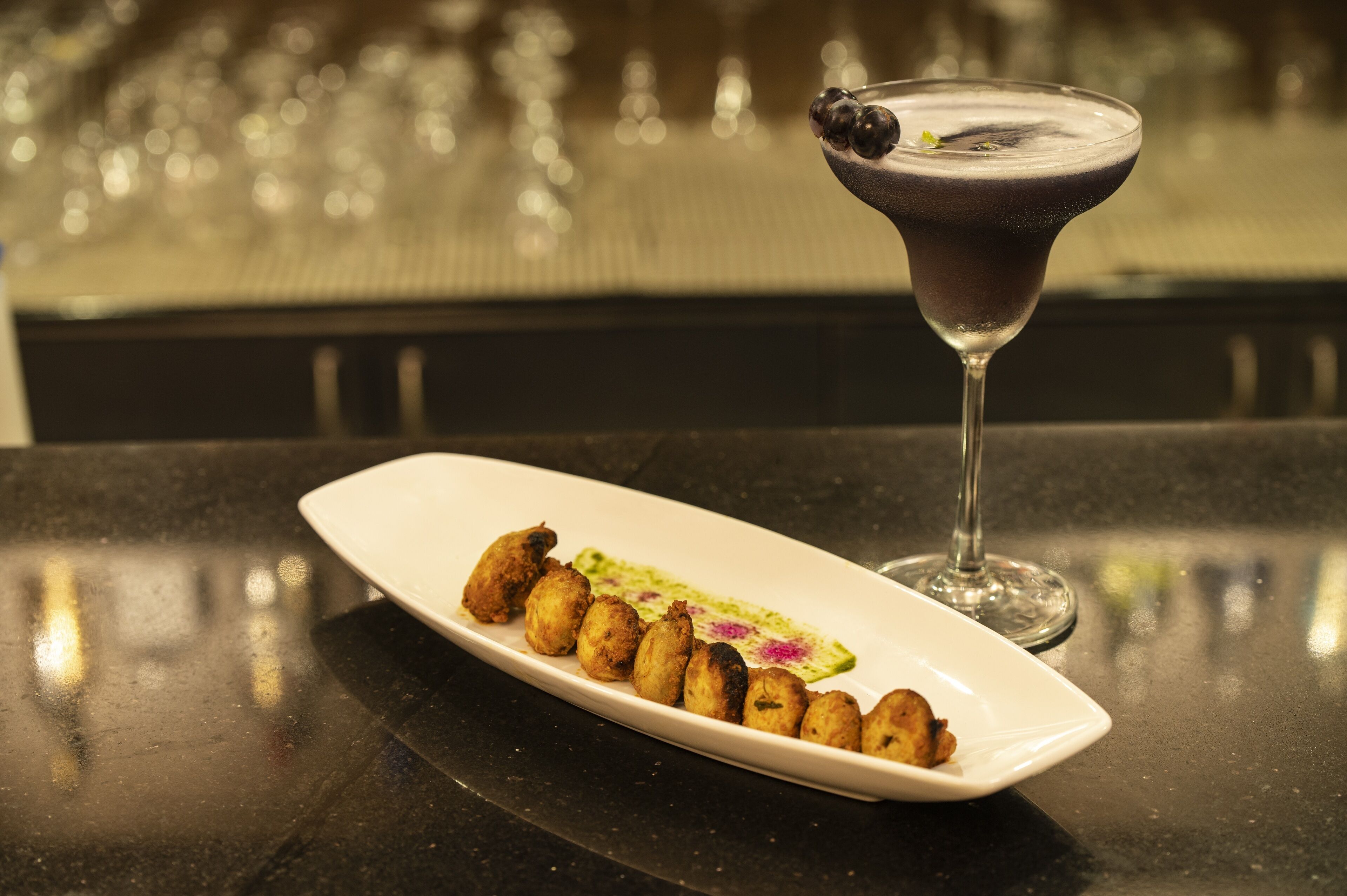 A white rectangular plate with fried items and a green garnish, accompanied by a dark-colored cocktail in a stemmed glass.