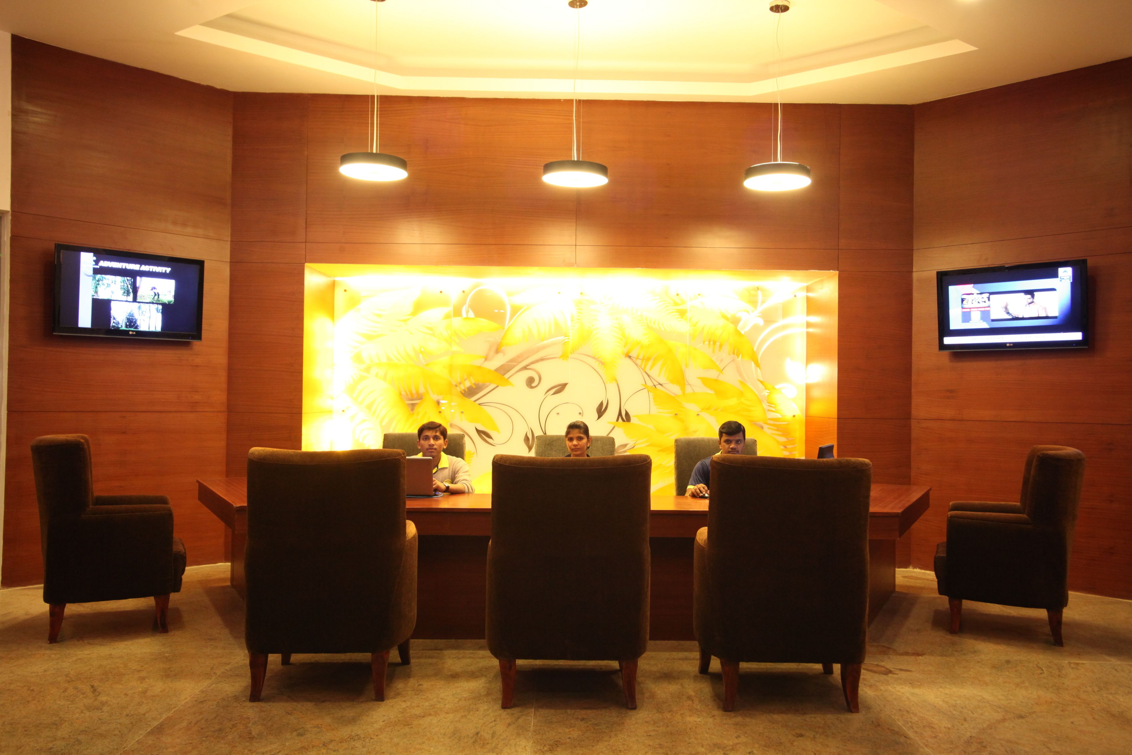 A conference room with a large table and four chairs, three people seated, and two screens on the walls.