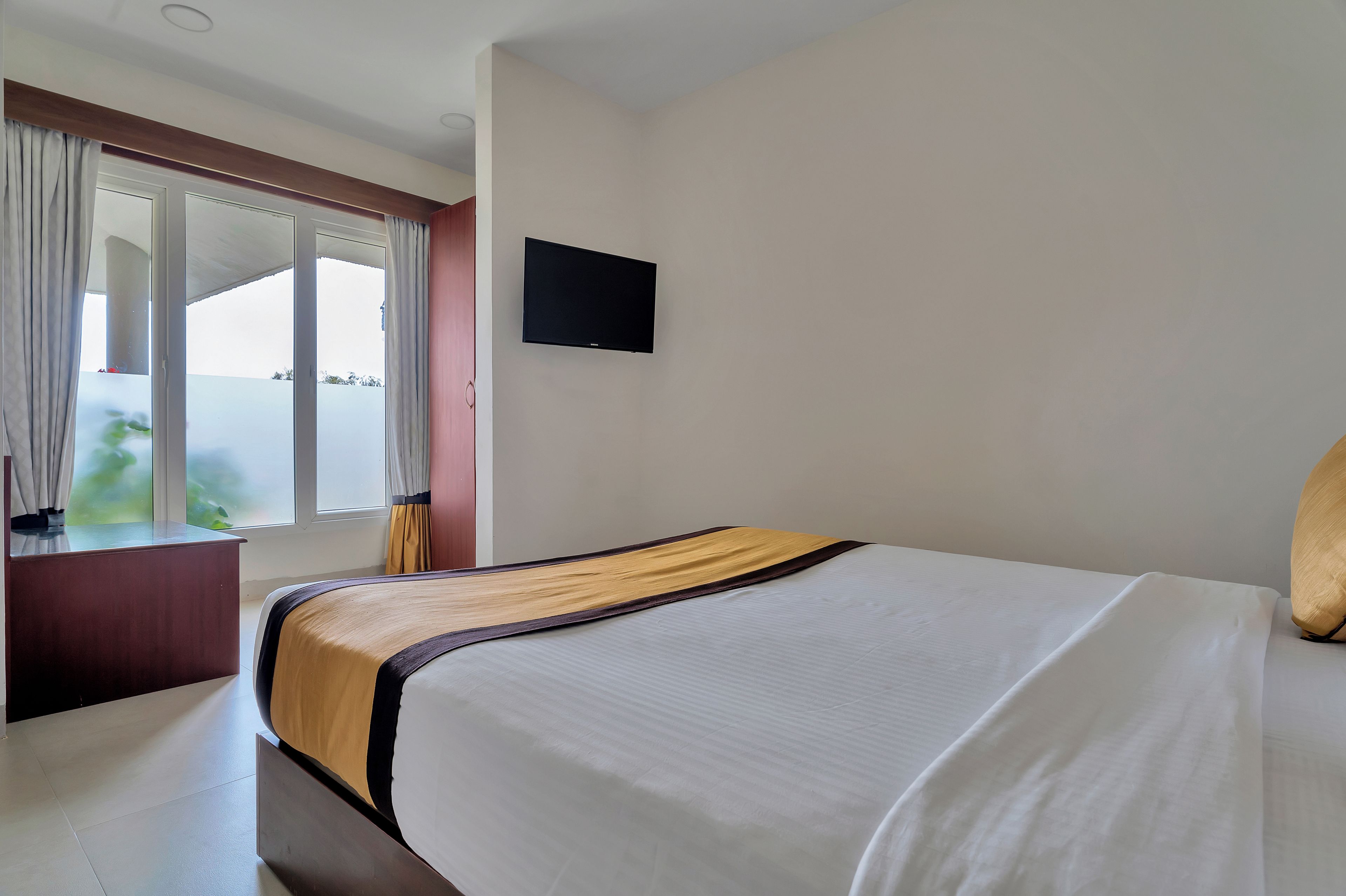 A hotel room with a bed, a television on the wall, and a window with curtains.